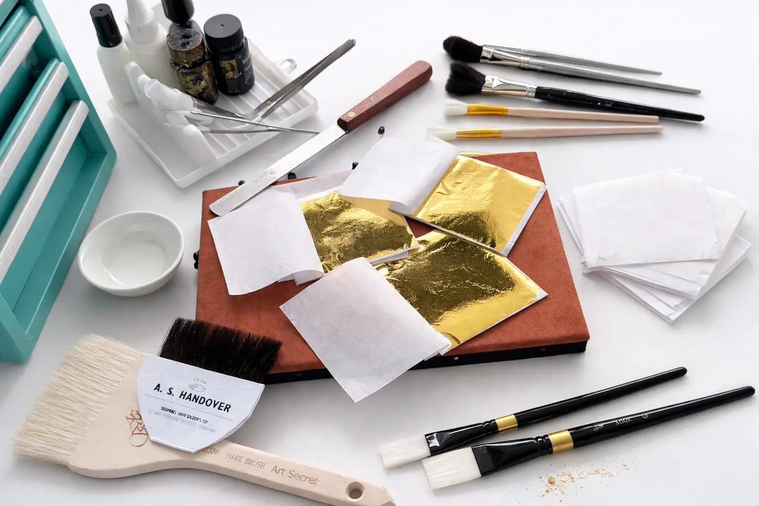Artist gold leaf tools and materials laid out for gilding, including loose gold leaf sheets, brushes, tweezers, adhesive, and a leather gilding cushion.