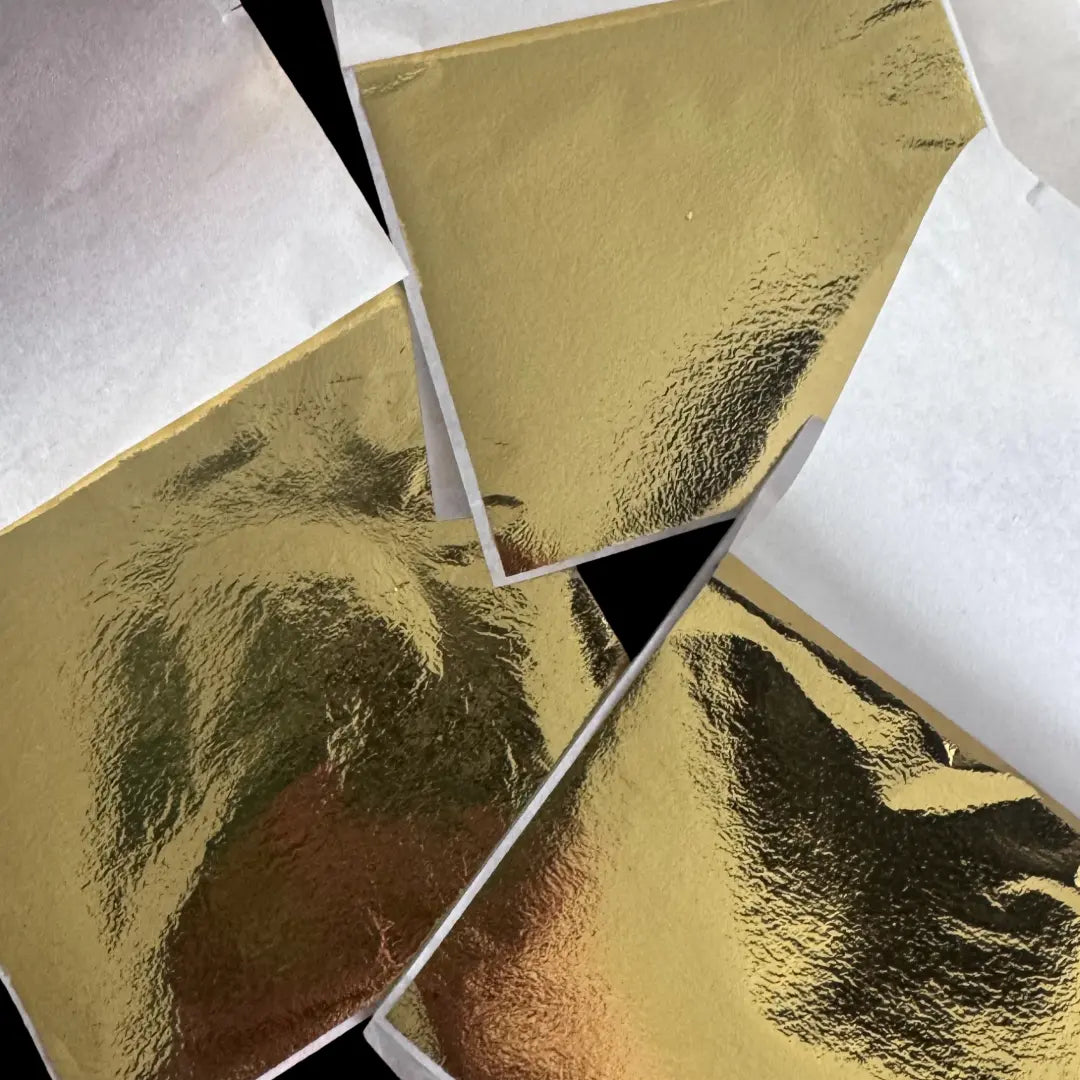 Three gold leaf sheets displayed on a neutral surface, showing traditional gilding materials.