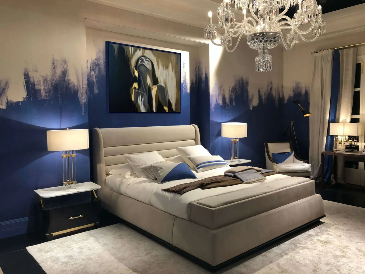 Release figurative mixed media artwork with 23K gold leaf displayed above a luxury bedroom featuring a large beige upholstered platform bed with white bedding and blue accent pillows, set against a dark blue and beige abstract wall. The room is styled with a crystal chandelier, bedside tables with lamps, and a matching armchair.