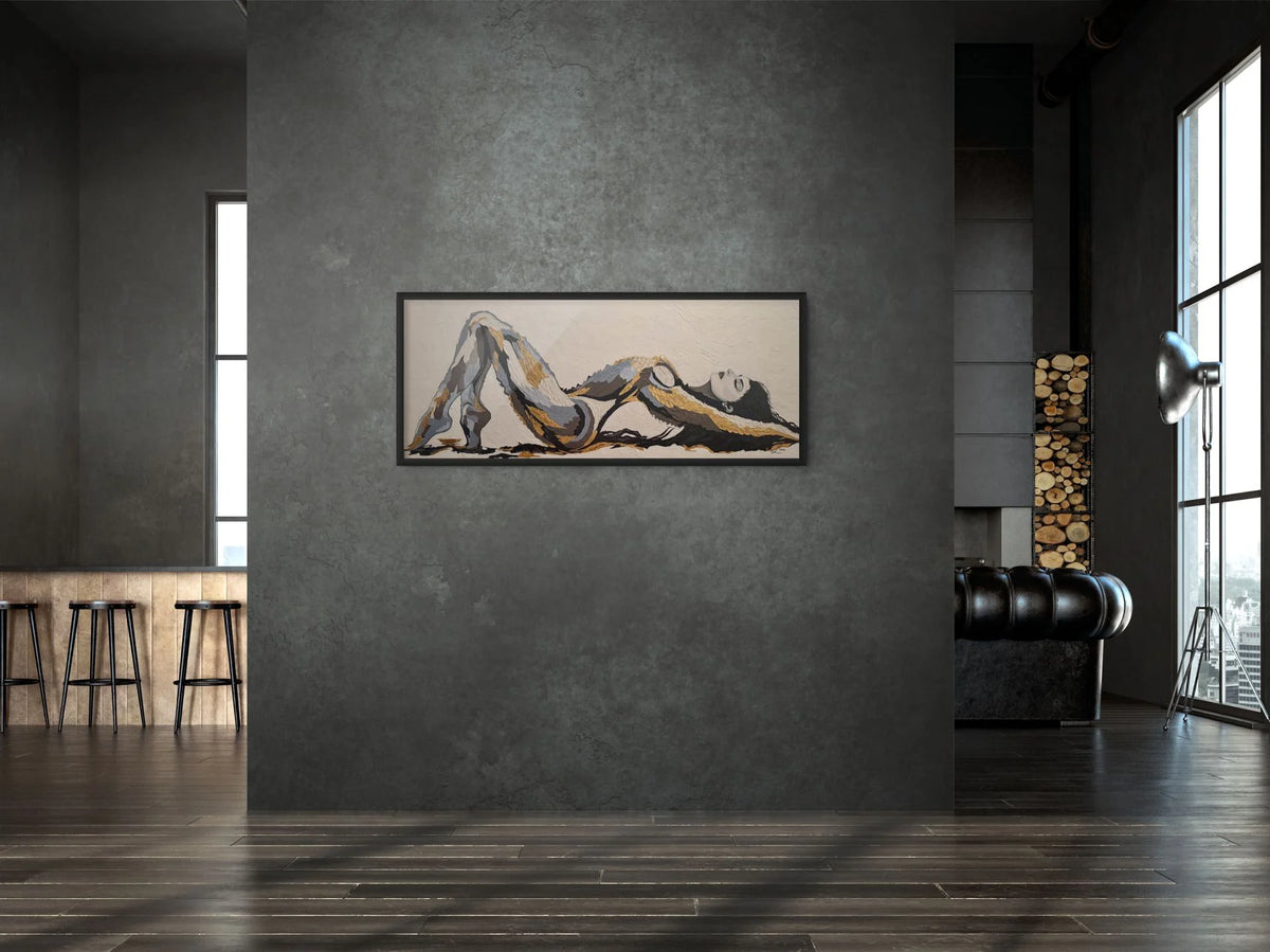 The Recline original mixed media figurative artwork with 23K gold leaf displayed in a contemporary interior with black frame. The painting features a stylised reclining female figure in beige, black, and grey tones, displayed on a dark grey textured wall in a modern loft-style room with dark wood floors and a black leather sofa.
